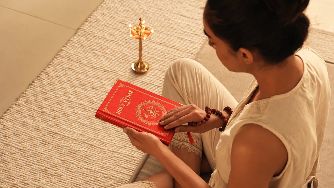 Vrouw die op een geweven mat het rode boek de Holy Vedas leest, naast een messing ayurvedische olielamp, voor Art of Vedas